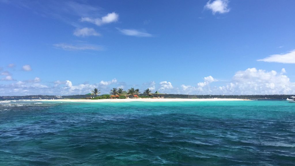 A white strip of beach, palm trees, and beach bars on Sandy Island in Anguilla seen from the sea