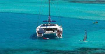 Wind surfers dart around a luxury catamaran floating in turquoise tropical waters
